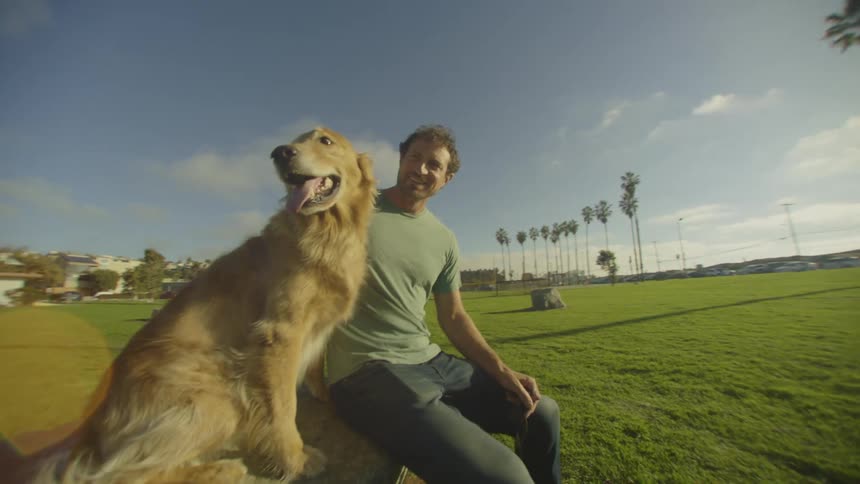 A young man sitting on a bench beside his golden retriever in a park at sunset smiling warmly as he gently pets the dog on its back while the soft orange light glows around them