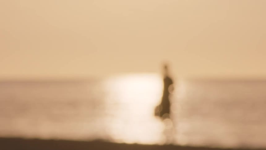 Person holding shoes on sand facing the sea at dusk