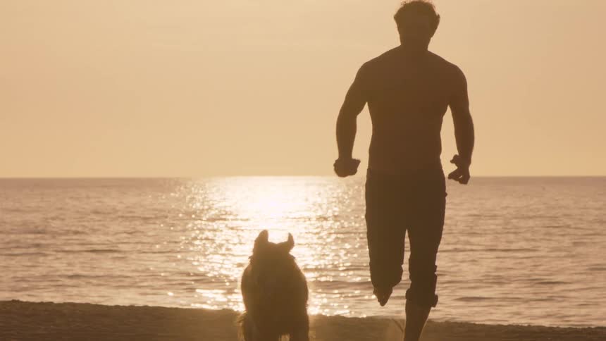 Man runs on the sand alongside a dog toward the sea at sunset