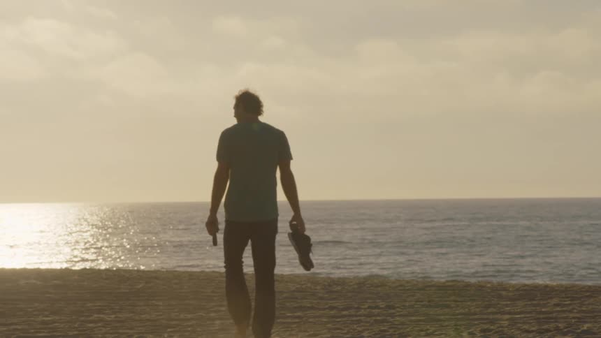 A man walks toward the ocean on a beach at sunset carrying his shoes