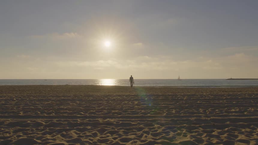 Man walking on the beach with a sailboat on the horizon as the sun shines over the water