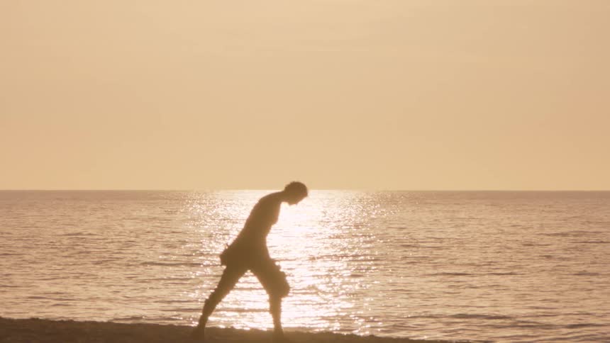 A mans silhouette pauses on a beach at sunset to kick sand into the water