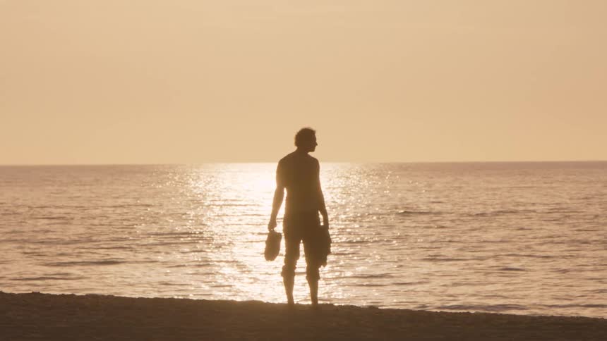A man stands in silhouette on a sandy beach during sunset looking out at the calm ocean while holding his shoes in his hands as the sunlight reflects off the waters surface