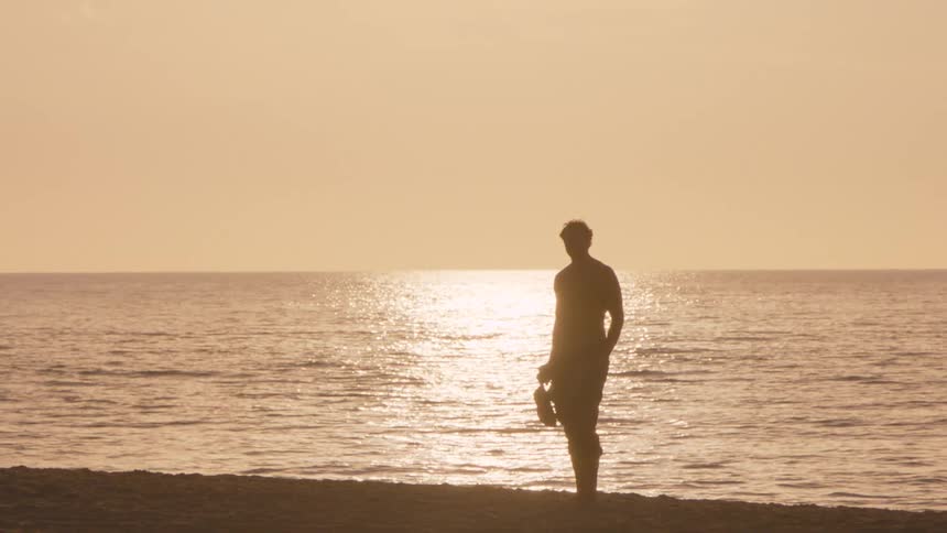 A man stands silhouetted on a sandy beach during sunset holding his shoes and clothes while looking out at the shimmering ocean waves as the camera slowly pulls into focus
