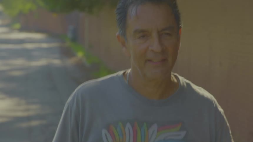 A middleaged man in a grey graphic tshirt walks toward the camera along a sunlit outdoor path next to a wall talking and looking around as he moves through the dappled light of the alleyway