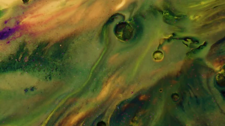 Vibrant shades of green gold and purple paint swirl together in a viscous liquid creating organic patterns and small craters as the fluids slowly merge and shift across the frame in a continuous macro shot