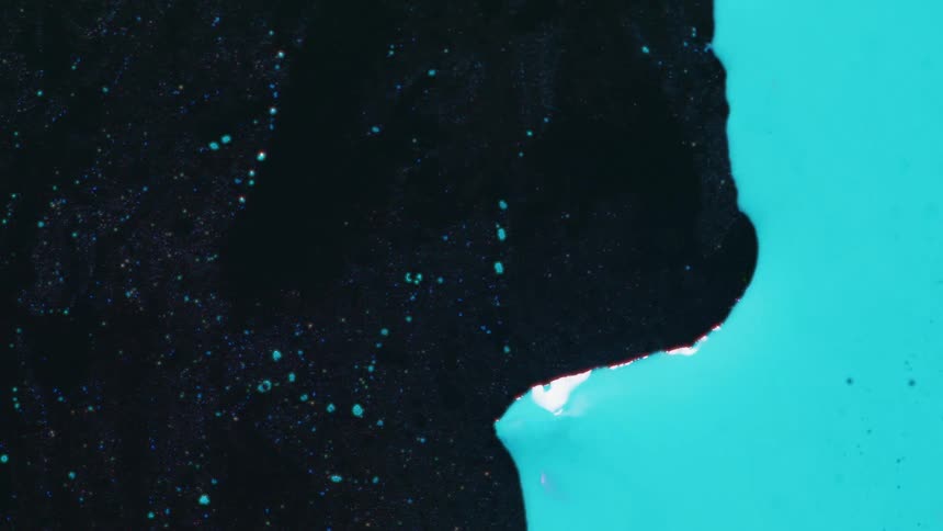 Opaque turquoise paint flows steadily across a dark textured surface covered in fine glitter eventually overtaking the frame and leaving behind small droplets and bubbles as the two viscous liquids merge and spread