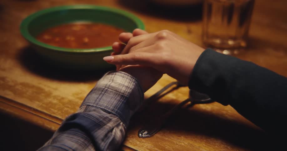 A pair of hands reaches out across a wooden dining table to clasp another persons hand beside a bowl of soupthe gentle gesture takes place in a dimly lit setting suggesting a moment of prayer comfort or connection before a meal