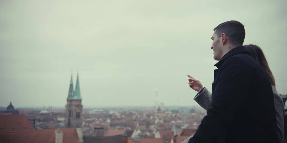 A couple stands on a balcony admiring the city view as the man points toward the horizon
