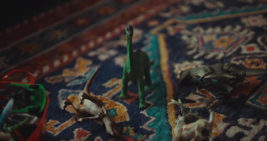 Toy dinosaurs including a tall green brachiosaurus and a pterodactyl are scattered across a patterned blue and orange rug next to a plastic toy basket in a dimly lit room