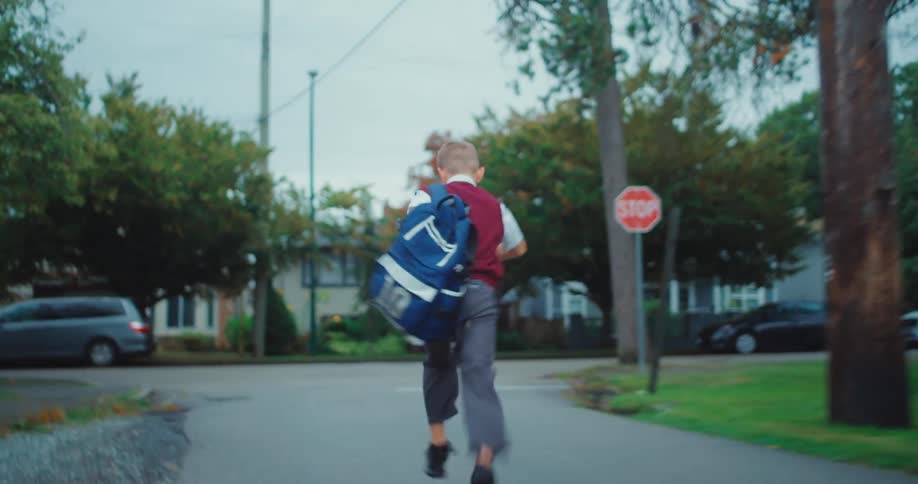 A young boy wearing a school uniform and a large blue backpack runs down a suburban streetthe camera follows him from behind as he heads toward a residential intersection with a stop sign
