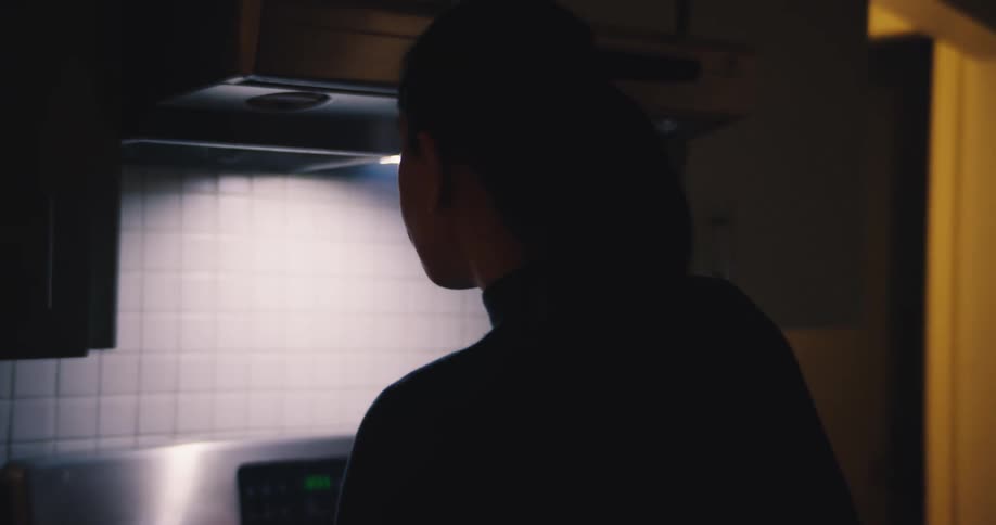 Young adult woman walking into a kitchen and pouring food from a pan on a stove into a ceramic bowl indoors during night time