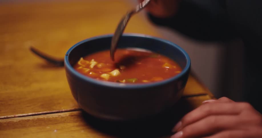 Adult female hands eating soup with a spoon from a blue ceramic bowl over a wooden table indoors
