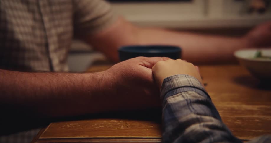 A man in a shortsleeved shirt and a person in a plaid shirt sit at a wooden table clasping hands for a moment of prayer before they release their grip and he picks up a spoon to begin eating from a blue bowl