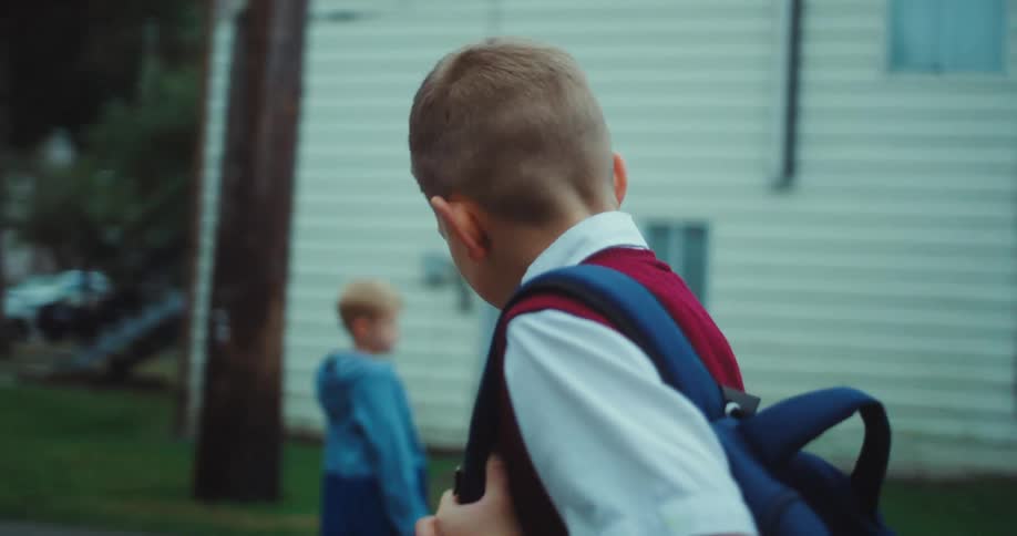 Young boy carrying a backpack walking with a sad facial expression next to other kids playing and laughing outdoors during day time