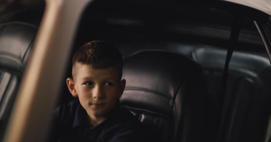 A young boy sits in the drivers seat of a car looking ahead and smiling before leaning out of the frame