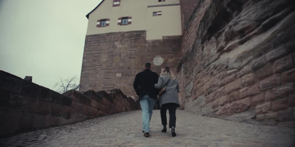 Couple holding hands walking alongside a rock wall with a building in the background