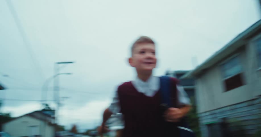 A young schoolboy in a maroon sweater vest runs excitedly down a residential street
