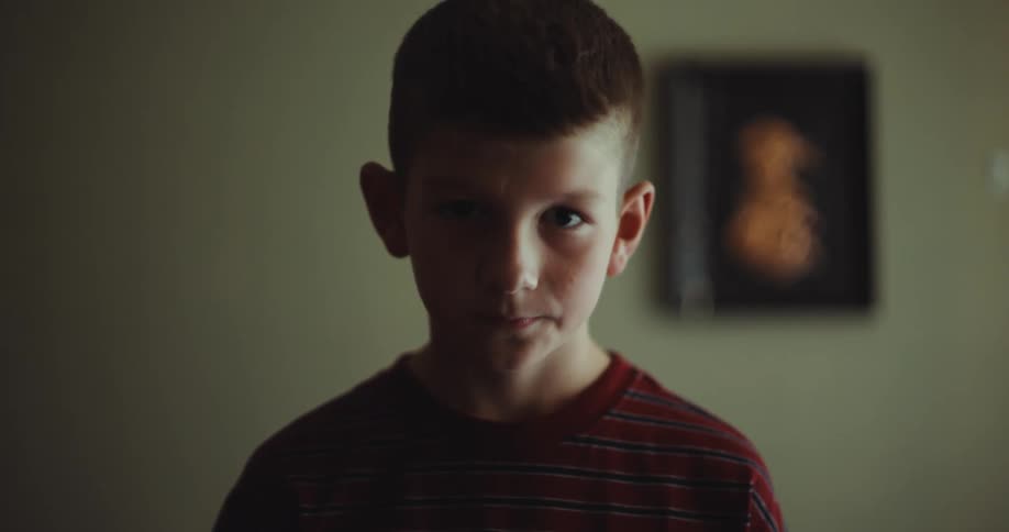 A young boy in a striped shirt stands in a dimly lit room looking directly into the camera with a neutral expression after briefly glancing downward he raises his eyes back toward the lens as the light shifts and fades across his face