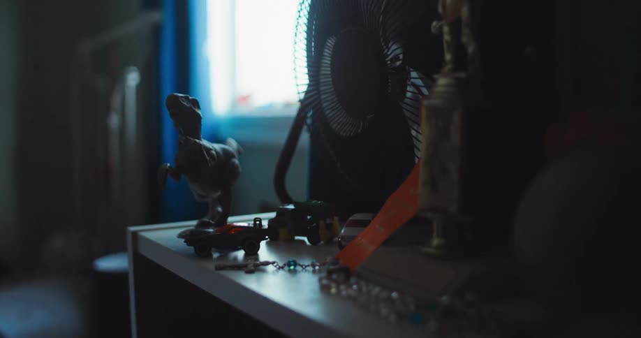A shelf beside a bedroom window holding a small fan and several toy figures including a dinosaur displayed among them