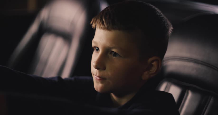 A young boy sits in a car looking forward thoughtfully before leaning out of the frame