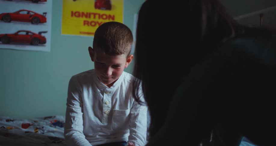 A young boy sitting on his bed at midday looking sad as his mother stands beside him gently talking to him in the soft natural light coming through the bedroom window