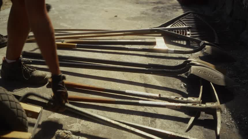 A worker places a crowbar onto a collection of shovels rakes and pickaxes lying on the ground