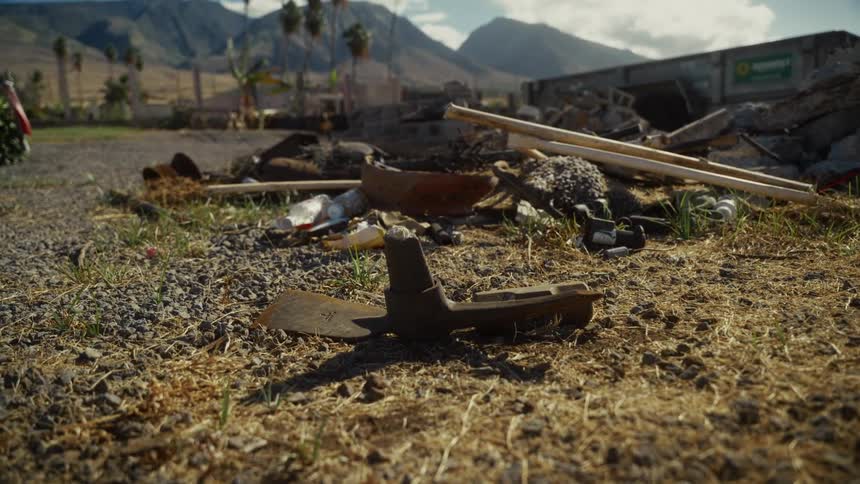 The video shows a closeup of a rusted garden tool head likely a hoe or pick resting on dry rocky ground in the background there is a pile of debris and rubble set against a landscape of mountains and palm trees under a clear sky