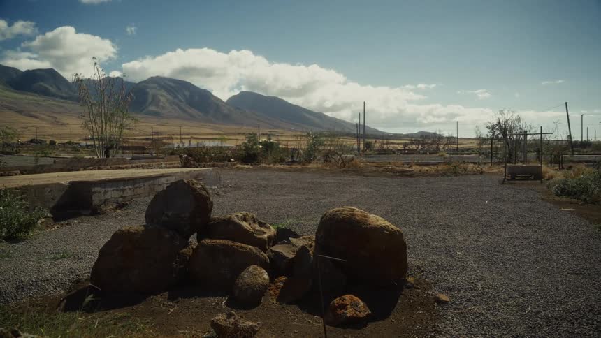 A wide stretch of barren countryside at midday the dry land scattered with rocks and patches of rough earth while a distant mountain range rises along the horizon under the bright overhead sun