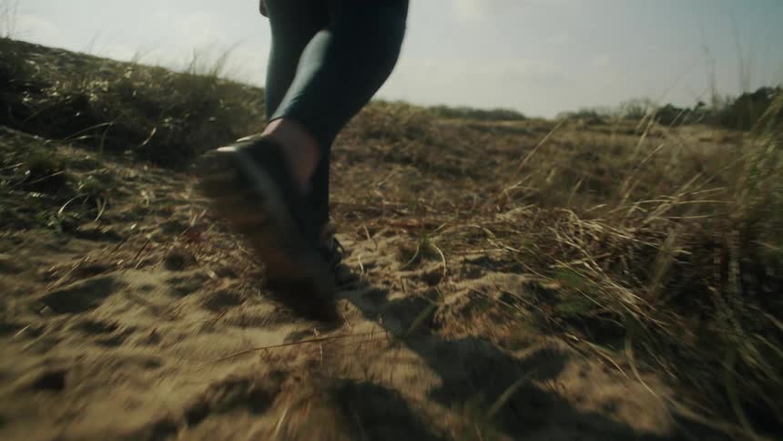 A person in athletic gear walks along a sandy sunlit path through dry grass