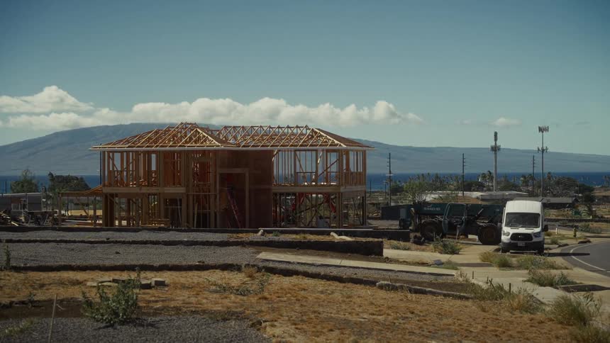A partially framed house stands under construction in a dry landscape with mountains and clouds in the distance