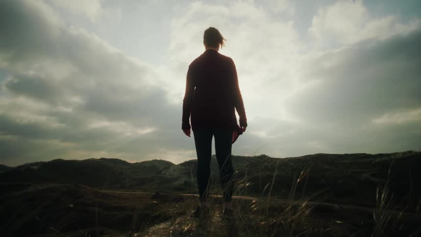 A silhouetted woman walks onto a grassy ridge to overlook a vast sun drenched landscape