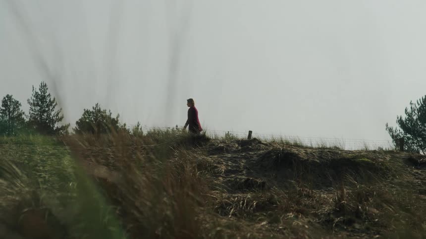 Woman walks along a dune covered with low vegetation with trees in the background under an overcast sky