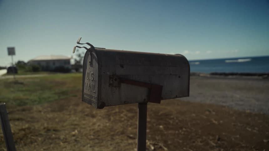 A weathered mailbox stands along a sunlit coastal road overlooking the sea