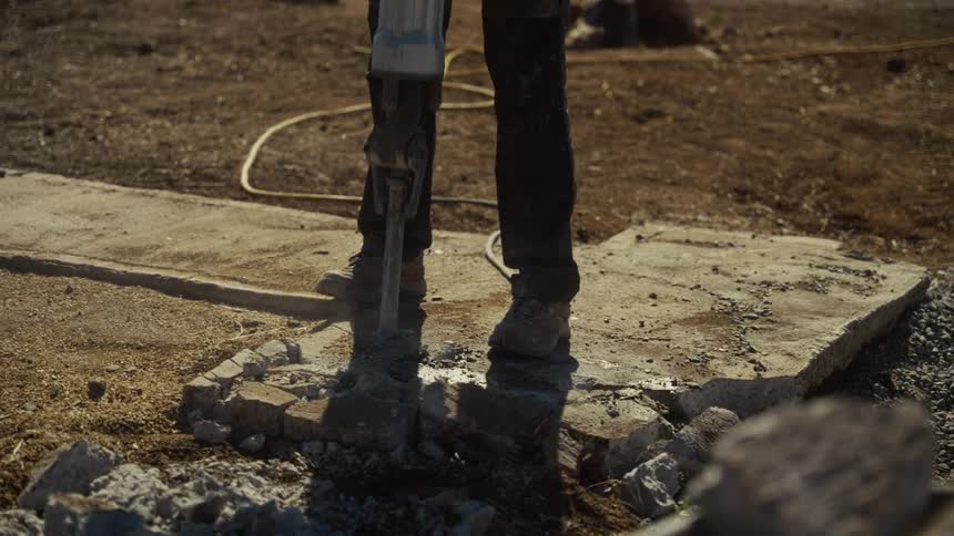 A construction worker uses a powerful jackhammer to break through a thick slab of concrete on a dusty outdoor site the tool vibrates intensely as it strikes the ground sending small pieces of debris and dust scattering across the uneven surface