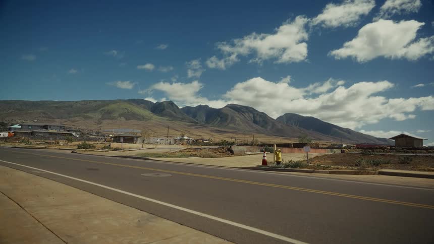 Paved road crosses a semiarid area with sparse buildings and a mountain range in the background under a partly cloudy sky