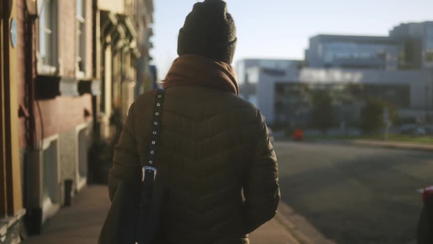 A young woman wearing a jacket and carrying a handbag walks along the sidewalk at midday sunlight casting clear shadows as she moves through the bright busy street