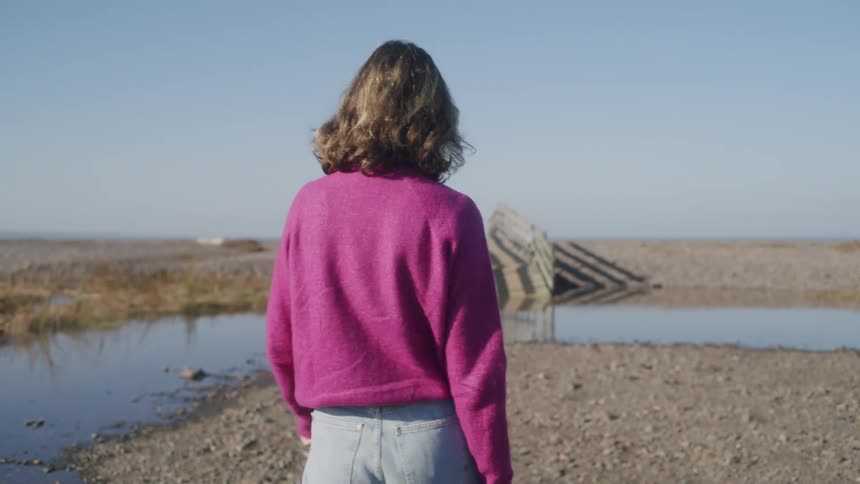 Woman walks away through a coastal area with shallow water pools and a wooden structure in the background