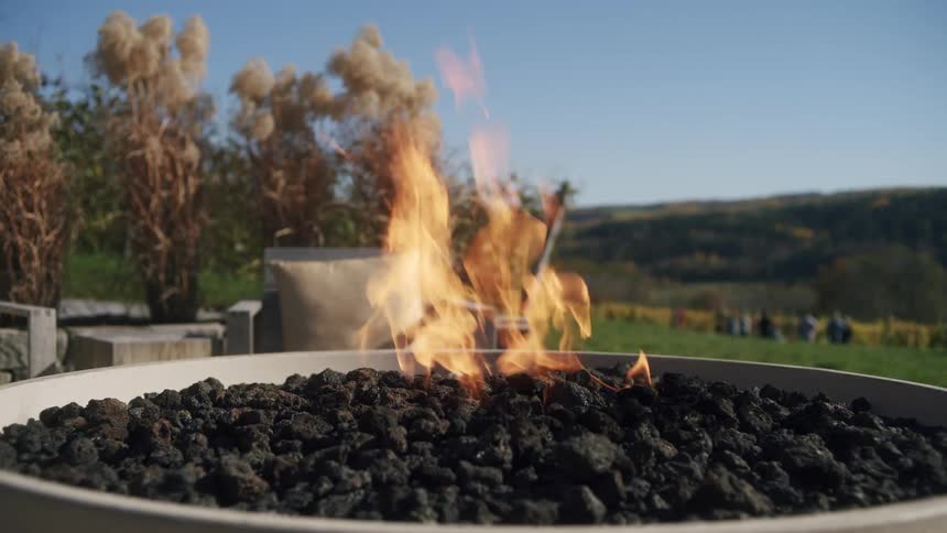 Controlled flame burns over volcanic stones in an outdoor area with a rural landscape in the background
