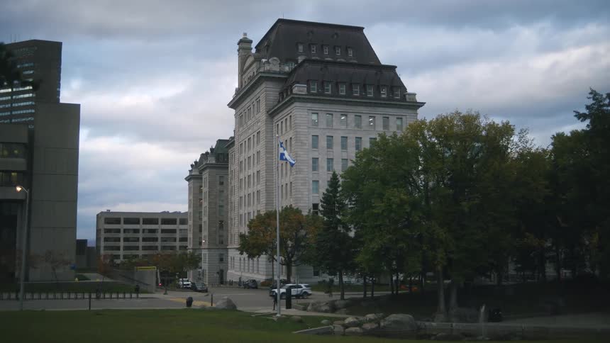 A large historic stone building with a dark mansard roof stands prominently under a cloudy sky in the foreground a blue and white flag flutters on a tall pole next to a street with light traffic and autumn trees