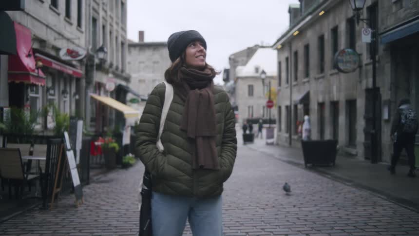Young woman stands on a downtown street smiling as she looks at buildings and a pigeon walks among pedestrians