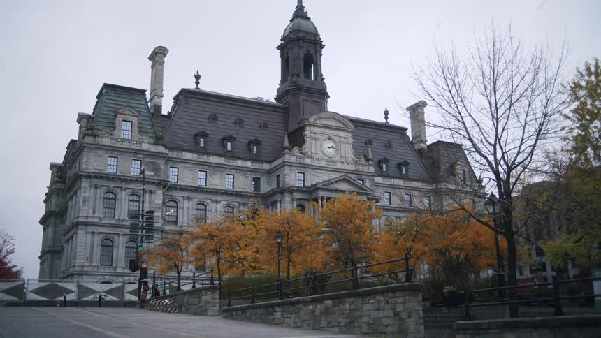 A view of montreal city hall on a cloudy day with a row of trees across the road displaying bright yellow fall leaves a car passes along the street in front
