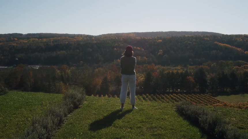 Woman stands on a grassy area overlooking a rural landscape with wooded hills in the background