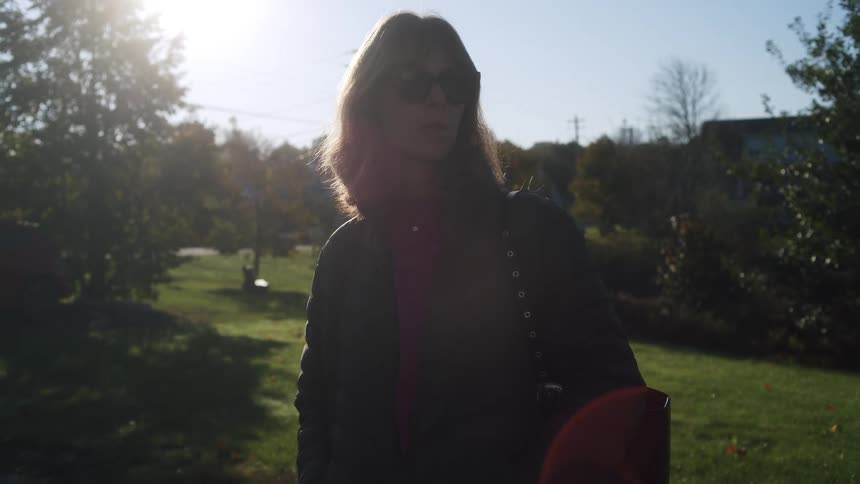 A woman wearing sunglasses and a dark puffer jacket stands outside in a sunny grassy yardshe looks off to the side while the bright sunlight creates a lens flare and highlights her hair from behind