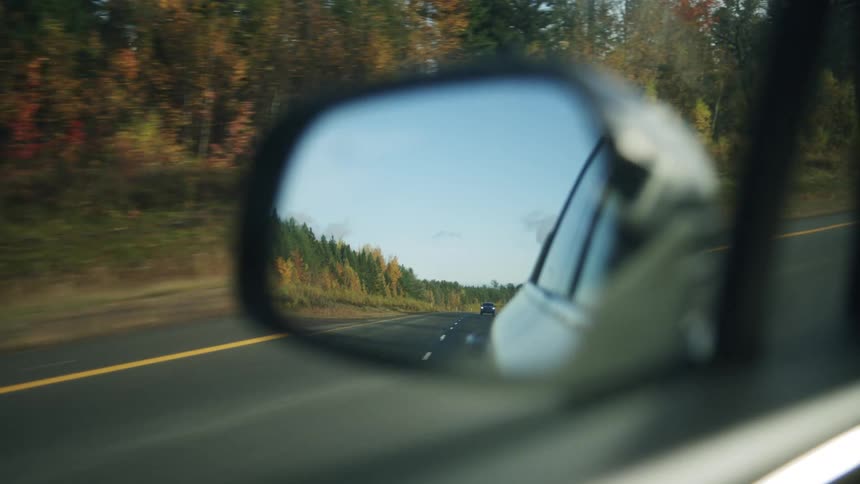 A drive through the countryside at midday with the cars side mirror reflecting another vehicle following behind the bright sunlight illuminating open fields and distant rural scenery on both sides of the road