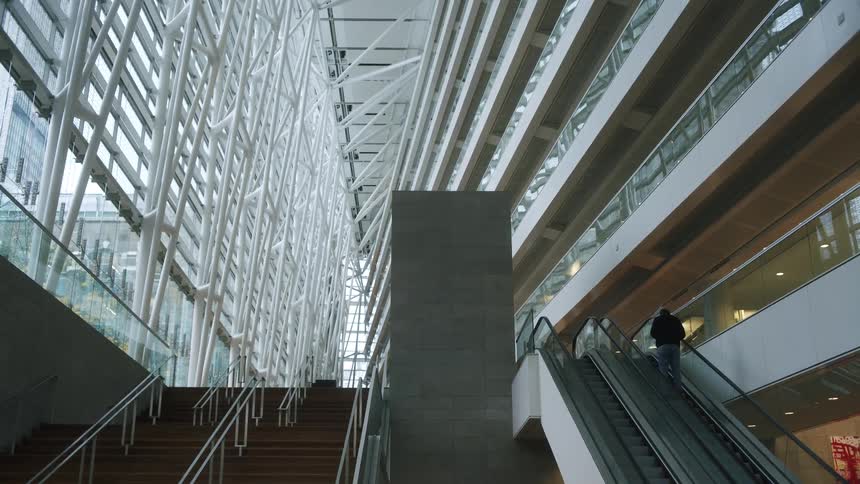 The video captures the interior of a modern multistory atrium featuring a complex white lattice structure along the glass facadeon the right side of the frame a person ascends an escalator past several floors of a brightly lit building