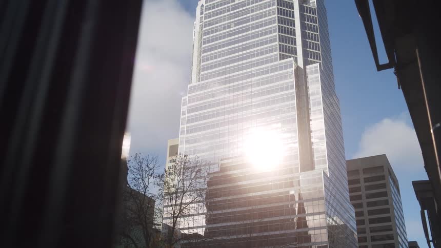 A tall modern glass skyscraper stands prominently in the center of the frame reflecting a bright burst of sunlight from its faceted surfaceto the left the dark traditional spire of a stone church with a visible clock face provides a sharp architect