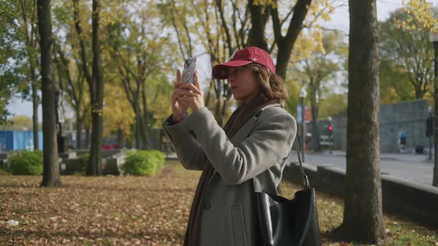 Young adult woman wearing a red cap and an overcoat taking pictures on her phone at a park in an urban area outdoors during day time