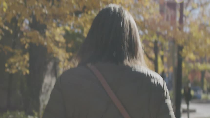 A woman with long dark hair walks away from the camera through a sunlit park or streetshe wears a dark quilted jacket and a brown crossbody bag strap while vibrant autumn leaves glow in the background