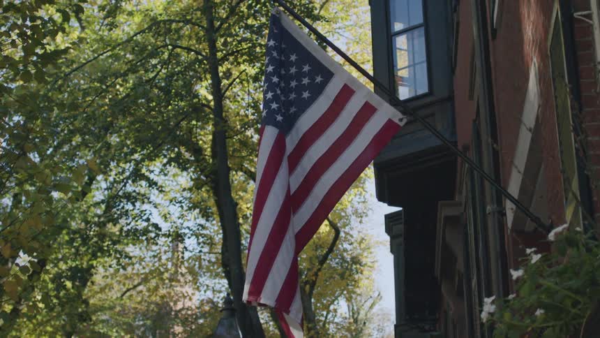 An american flag hangs from a black pole attached to a red brick building gently swaying in a light breezethe sun filters through the leaves of nearby green trees casting soft light and shadows across the flag and the side of the house